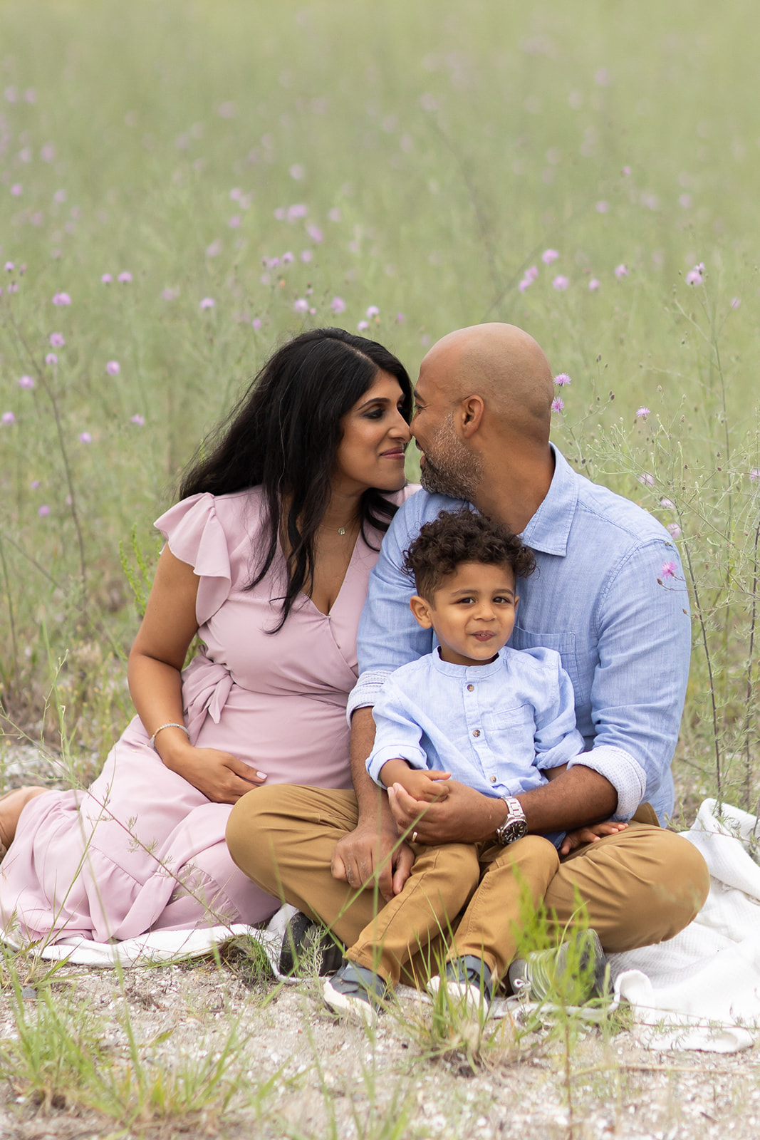 A mom and dad sit on a blanket in a field of wildflowers at a beach park with their son in dad's lap before visiting a Fairfield Public Pool