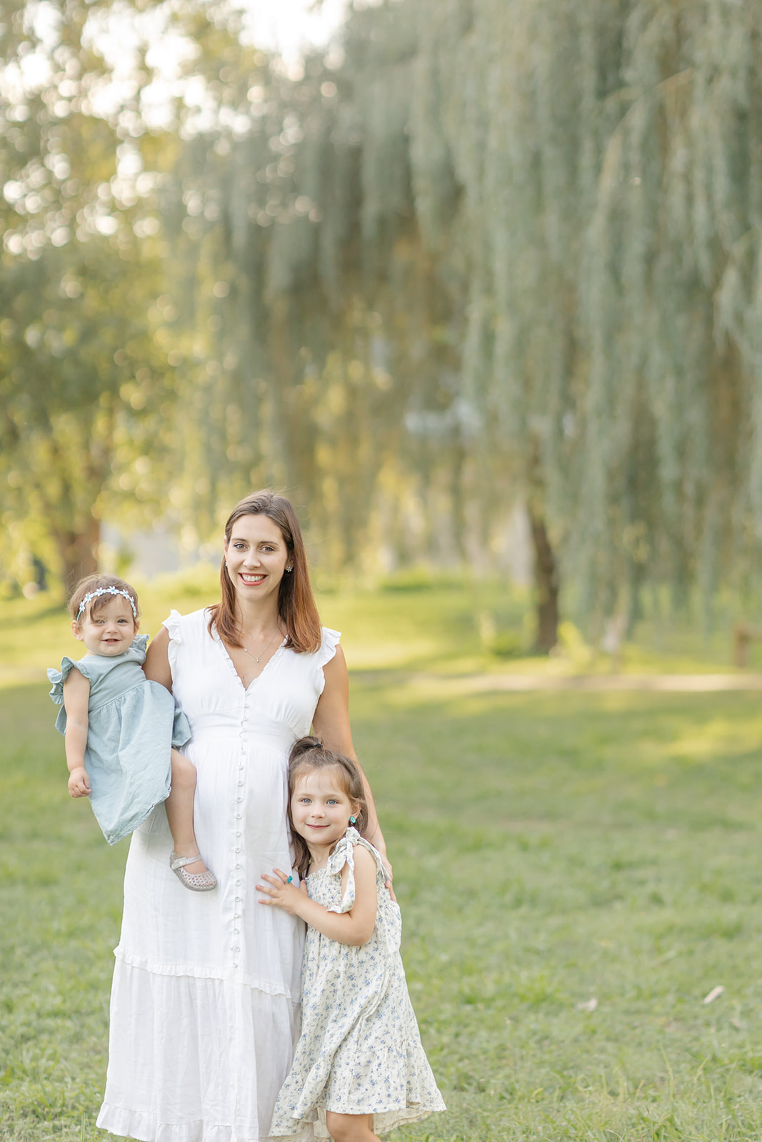 A pregnant mother stands in a park with her toddler daughter on her hip and older daughter hugging her leg