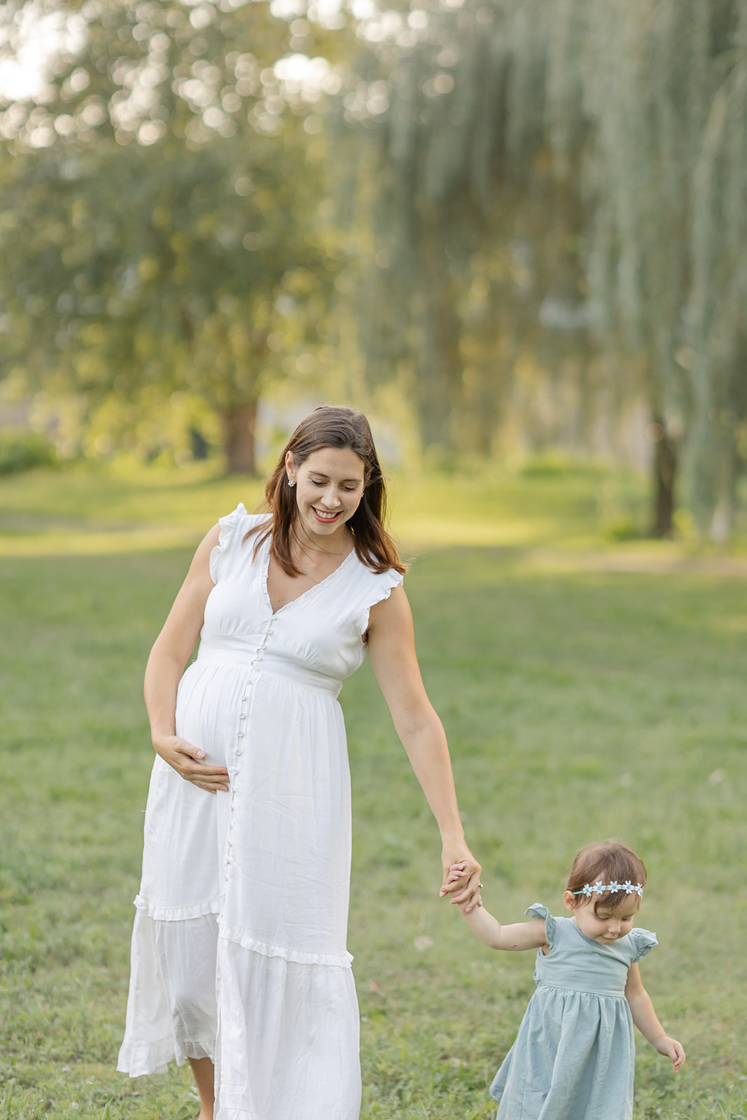 A pregnant mother holds her bump while walking through a park with her toddler daughter in a green dress thanks to Illume Fertility