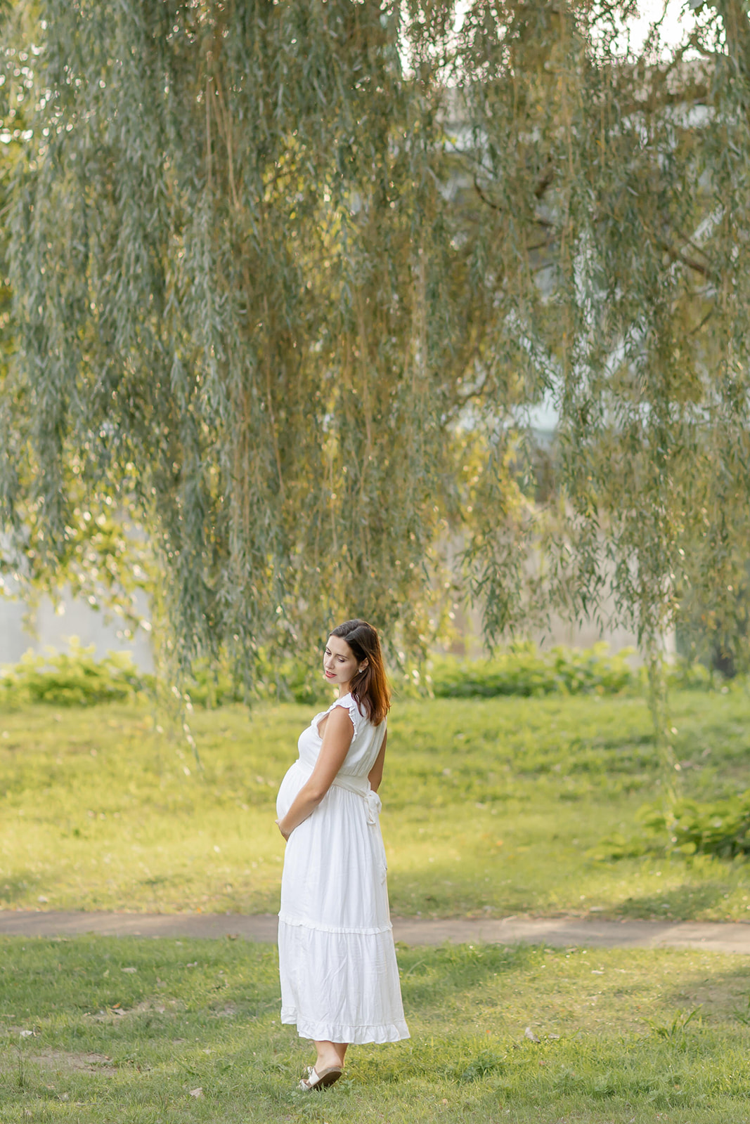 A mom to be walks under a willow tree at sunset in a white maternity gown thanks to Illume Fertility