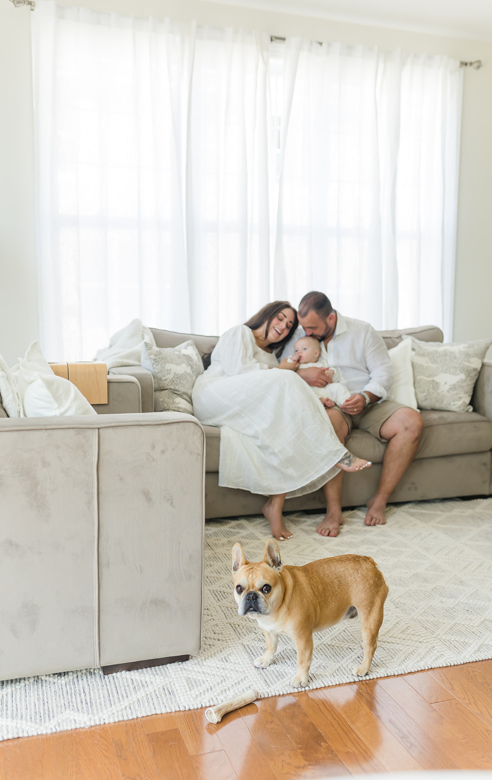 A mom and dad cuddle on the couch with their infant child while their dog stands in front of them on the rug