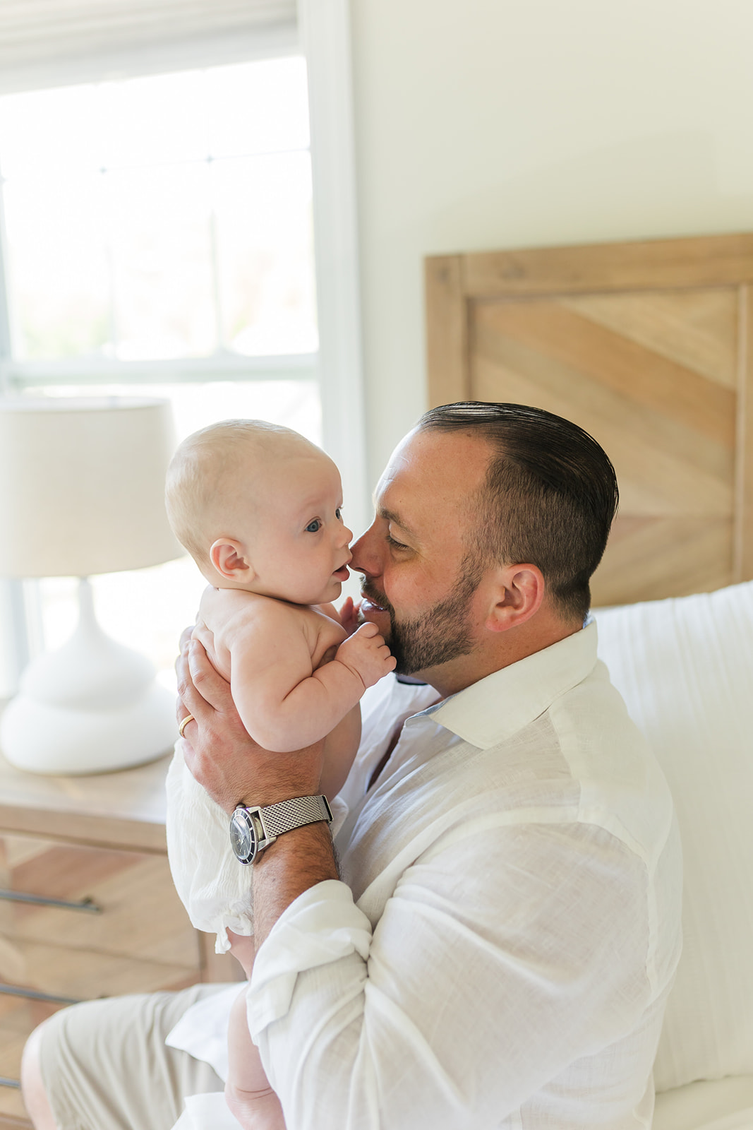 A dad plays and nuzzles his infant baby while sitting on a bed in a white shirt before visiting an Indoor Playground Fairfield CT