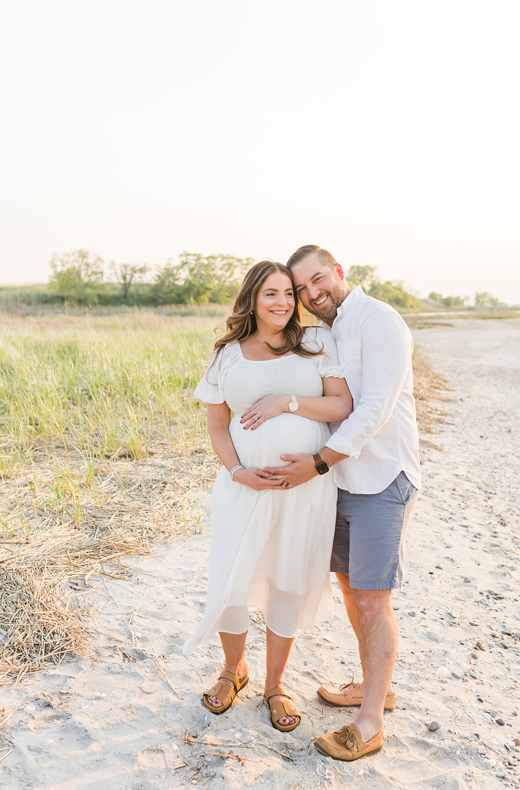 Parents to be smile while hugging on a beach and holding the bump at sunset