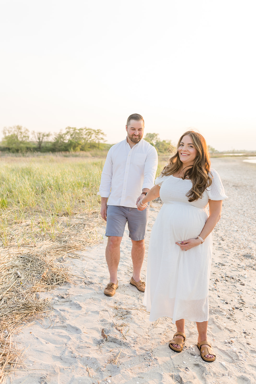 A mom to be in a white maternity dress walks down a beach holding hands with her husband at sunset after using IVF Connecticut