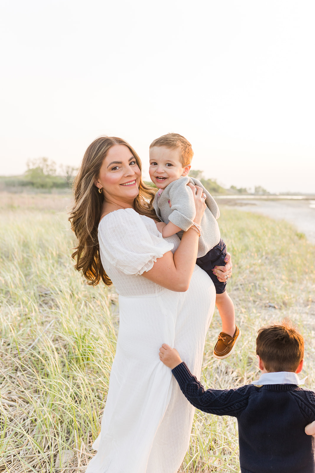 A pregnant mother in a white maternity dress plays on a beach with her two toddler sons at sunset after using IVF Connecticut