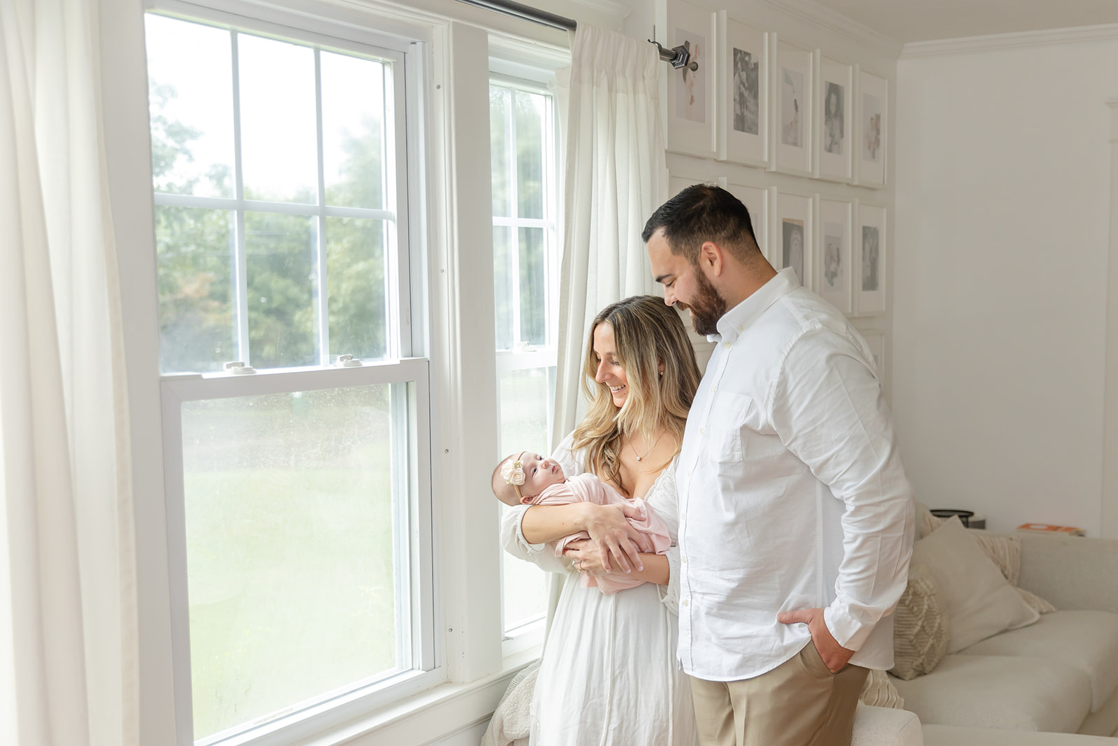 A mom and dad stand in a window with their newborn baby daughter in mom's arms