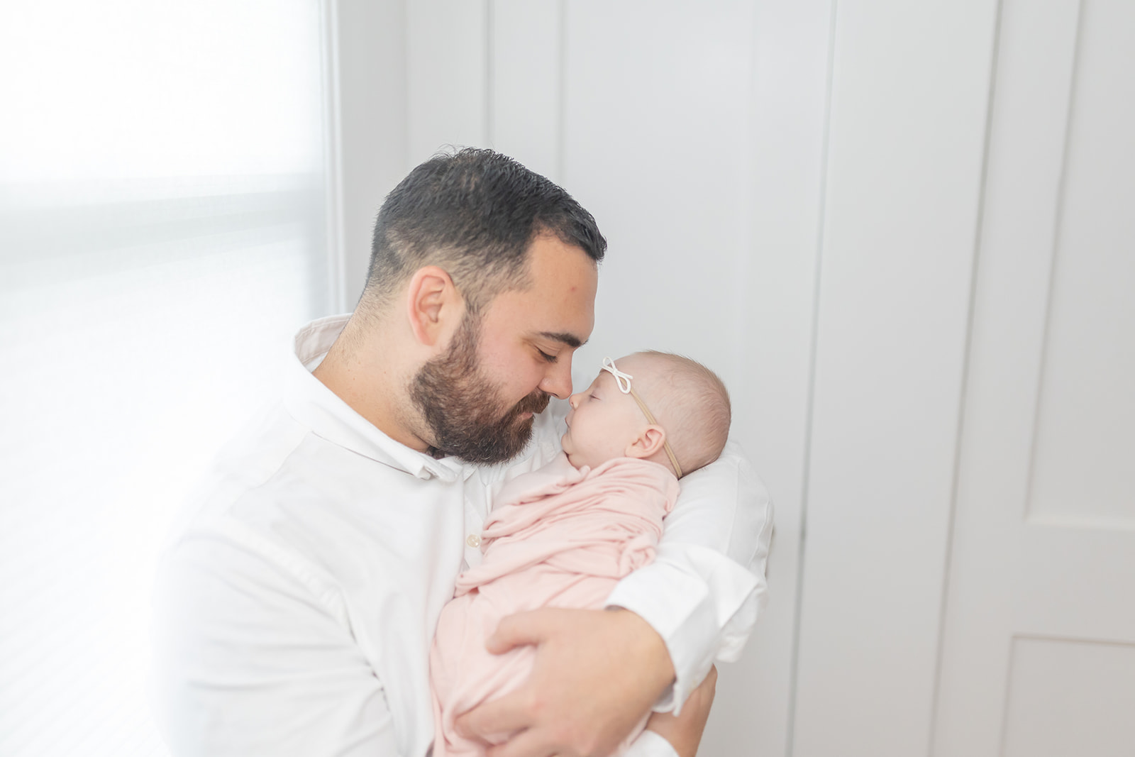 A father snuggles with his sleeping newborn baby daughter while standing in a window