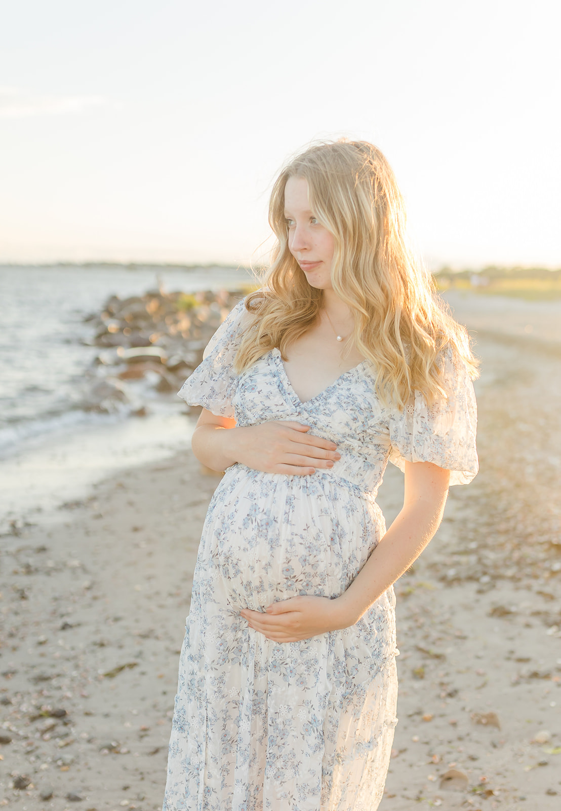 A mom to be stands on a beach at sunset holding her bump