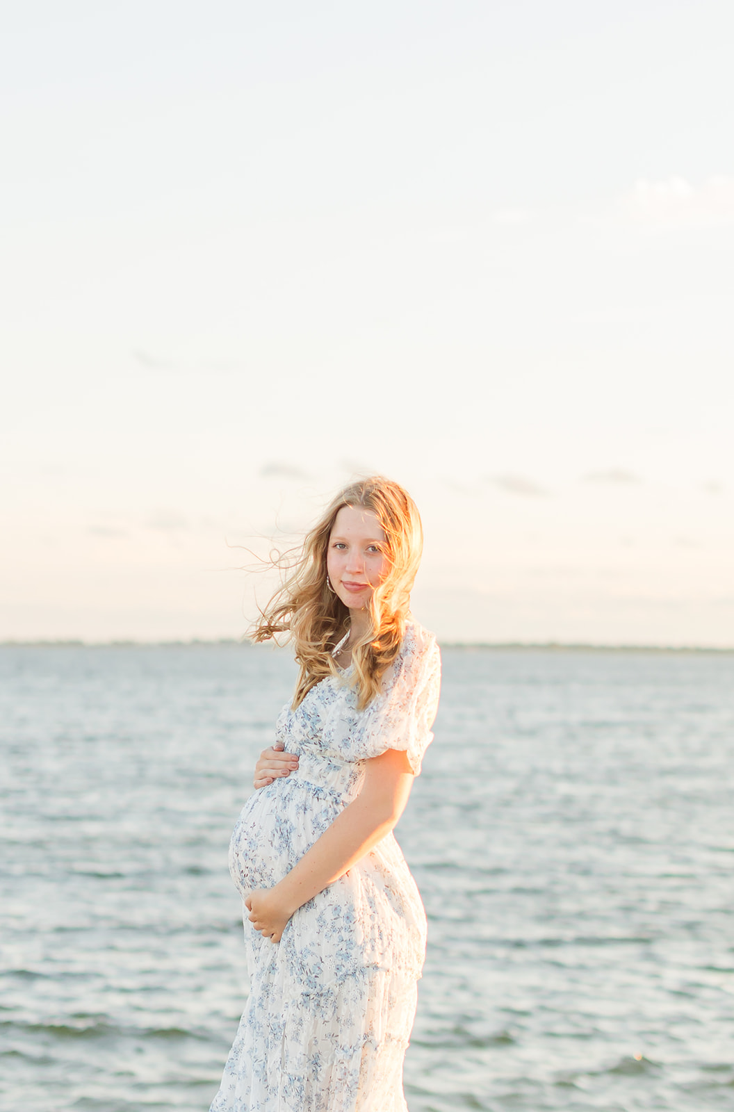 A mom to be smiles over her shoulder on a windy beach in a floral dress holding her bump after finding an OBGYN Bridgeport CT