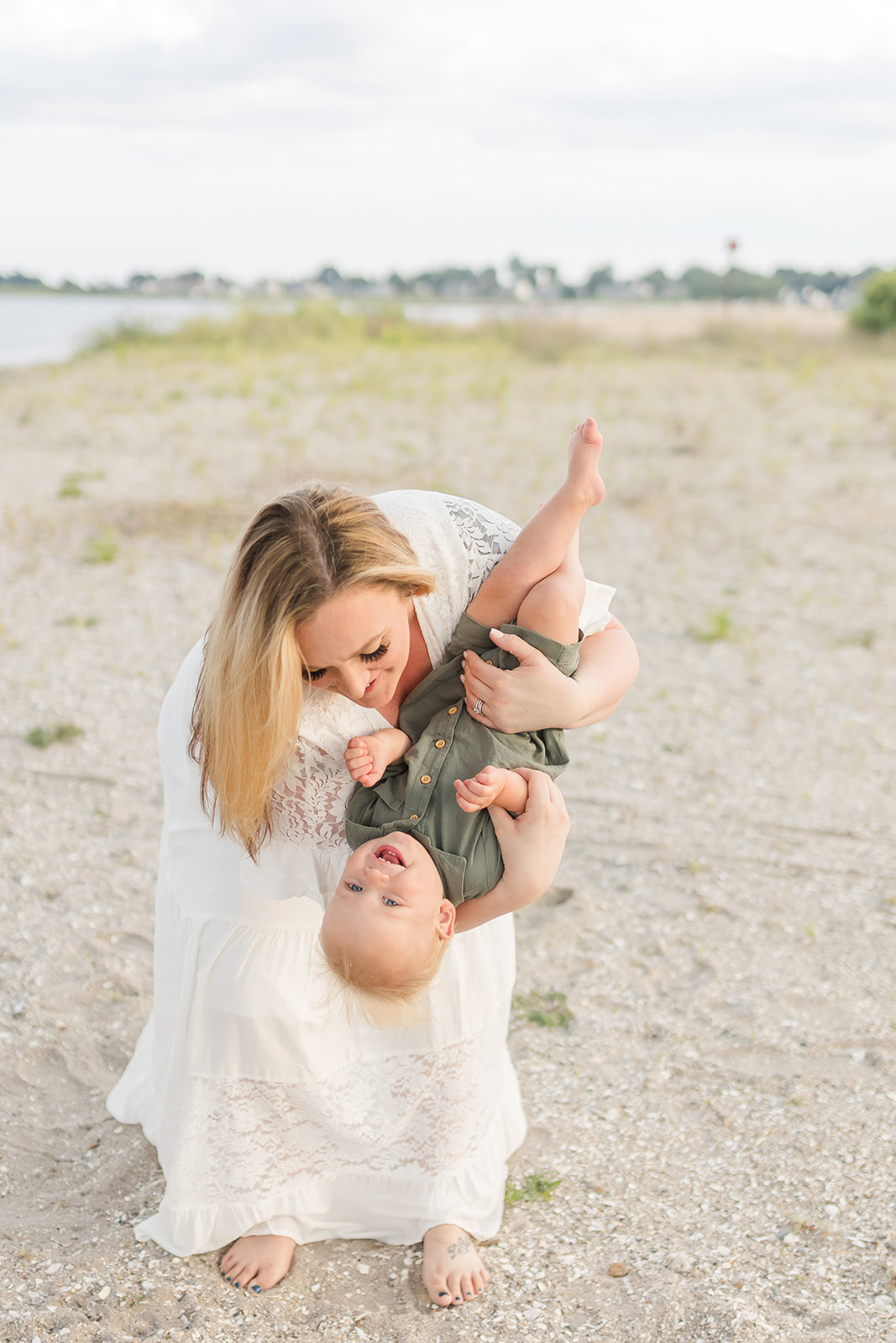 A pregnant mother in a white lace dress tips her toddler son while standing on a beach after visiting an OBGYN Fairfield County