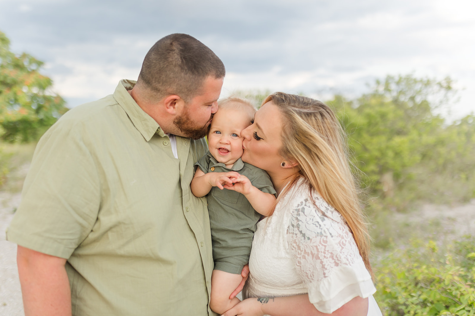 A mom and dad kiss the cheeks of their toddler son sitting in dad's arms