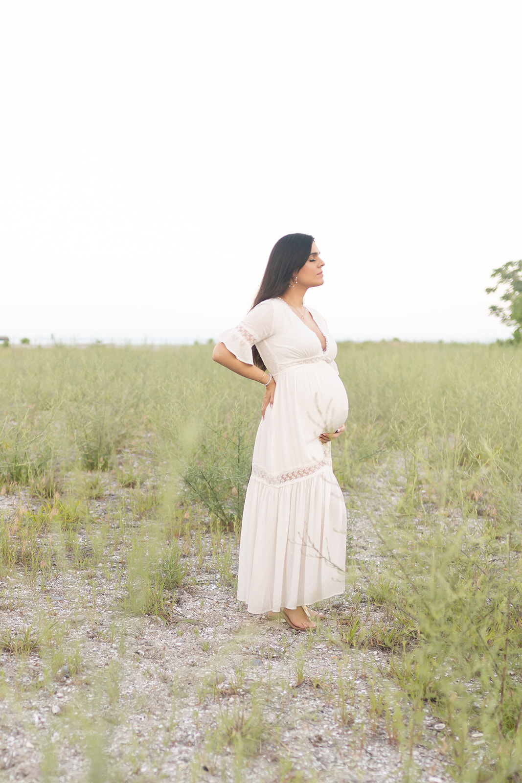 A mom to be stands in a field holding her bump in a white maternity dress after meeting an OBGYN Trumbull CT