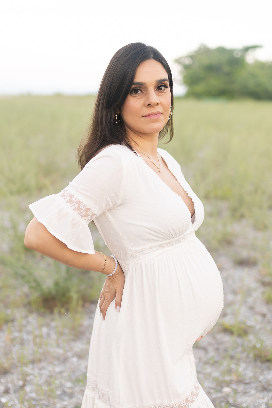 A mother to be stands in a field in a white maternity dress after meeting an OBGYN Trumbull CT