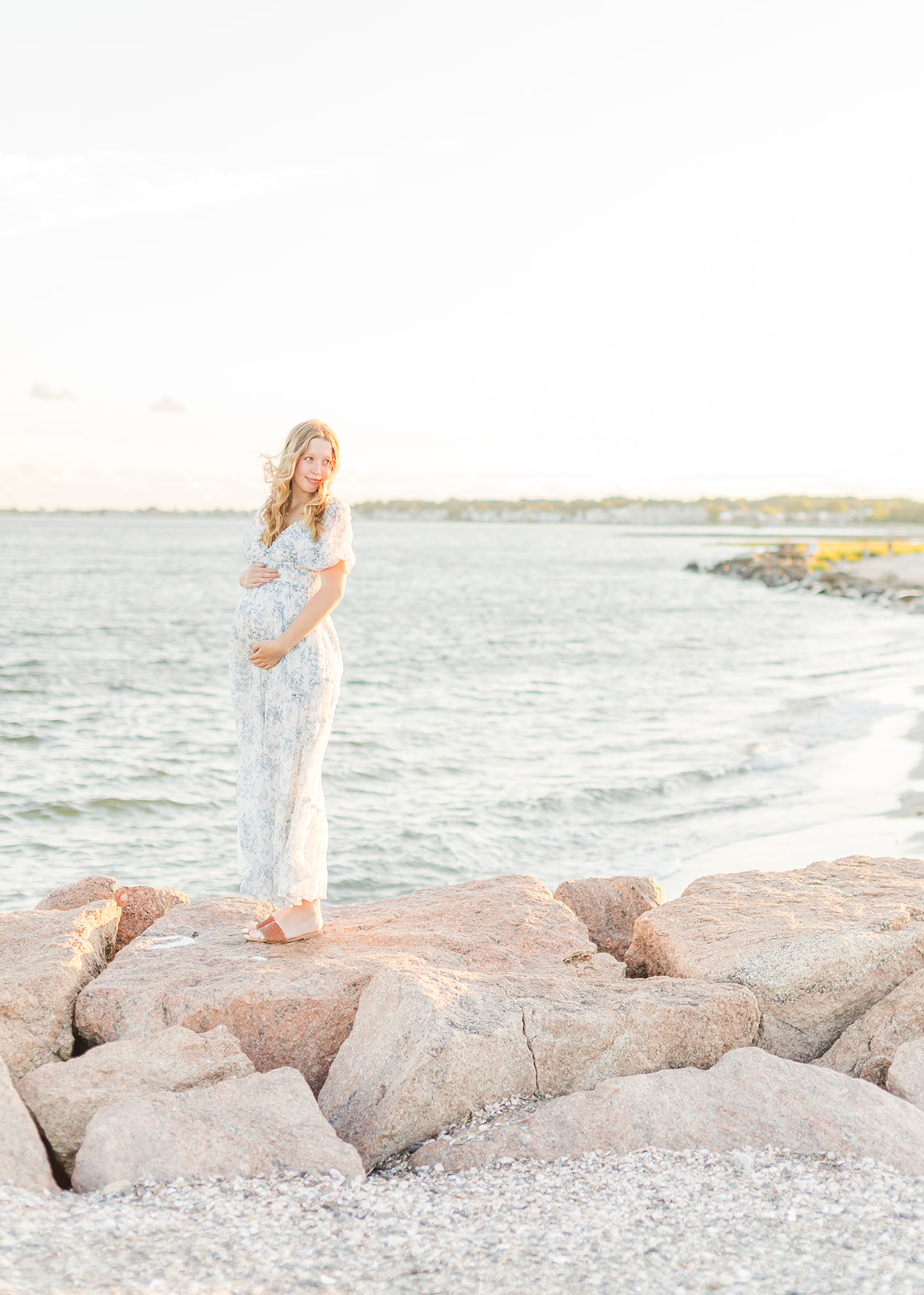 A mom to be stands on a rock seawall at the beach at sunset while holding her bump