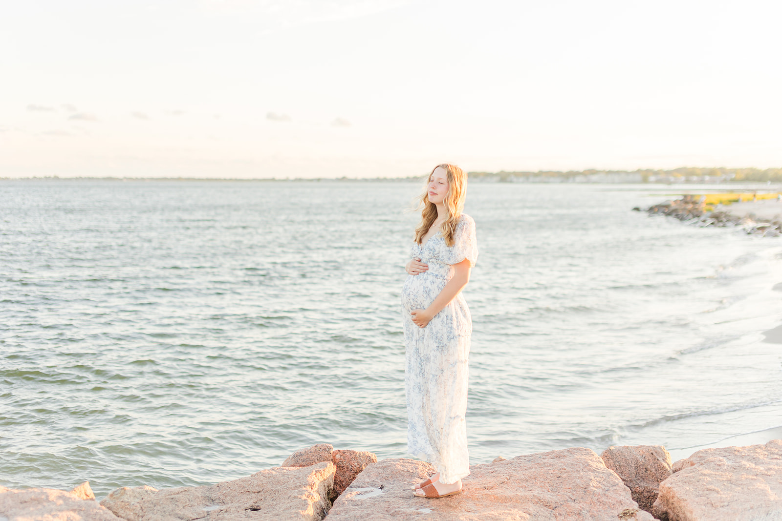 A mom to be holds her bump while standing on a beach rock with eyes closed thanks to Park Avenue Fertility
