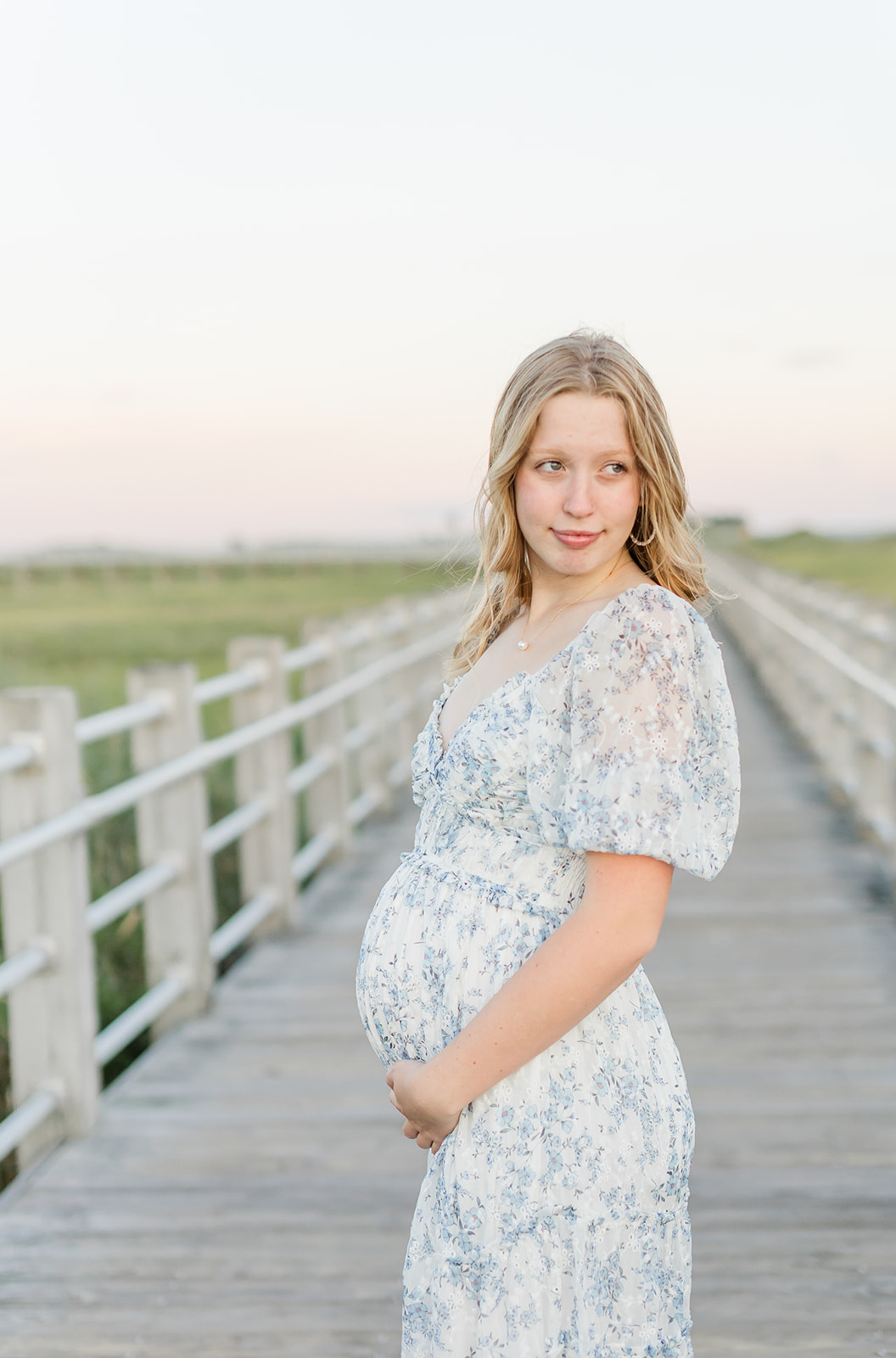 A mom to be in a blue floral dress looks over her shoulder while standing on a beach boardwalk thanks to Park Avenue Fertility