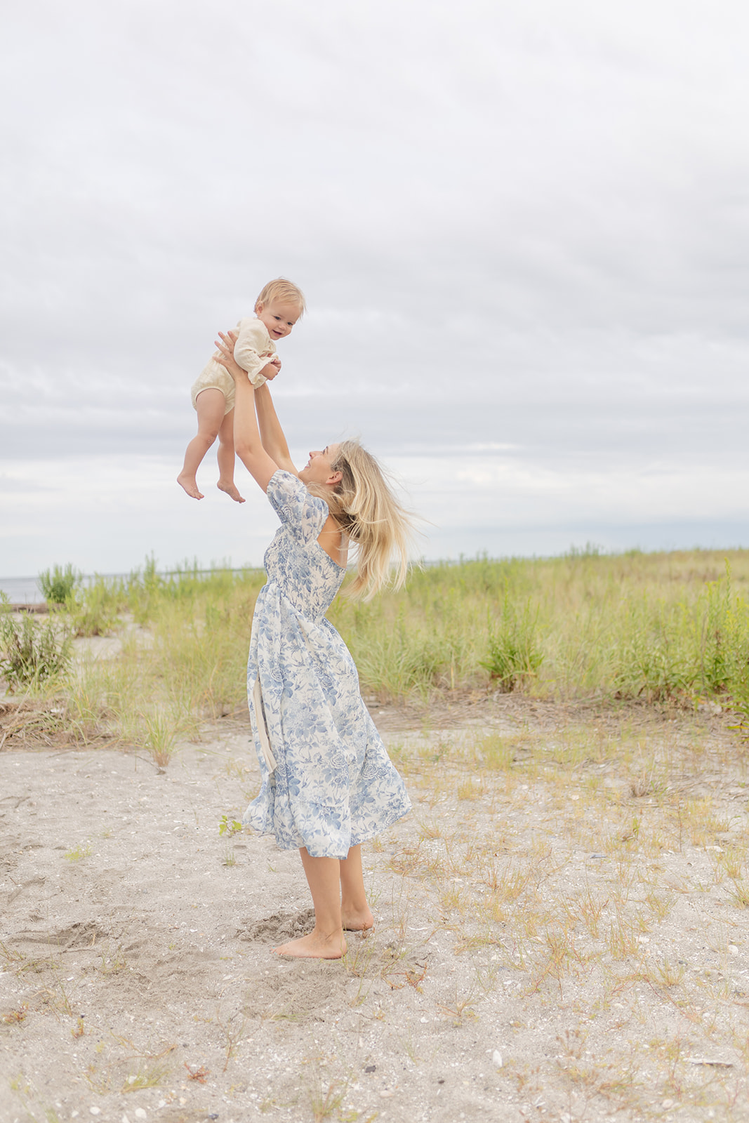 A mother in a blue floral dress tosses her toddler in the air on a windy beach