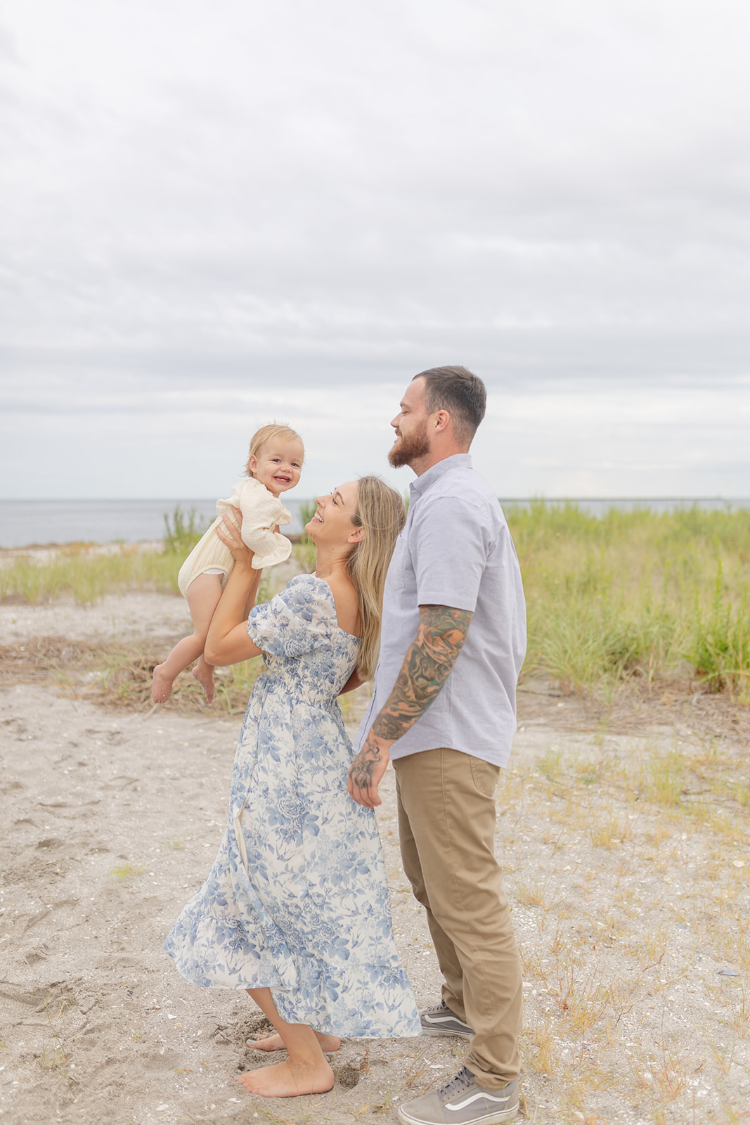 A mother in a blue floral dress plays and lifts her toddler daughter on a beach with dad behind her after visiting Playgrounds in Fairfield CT