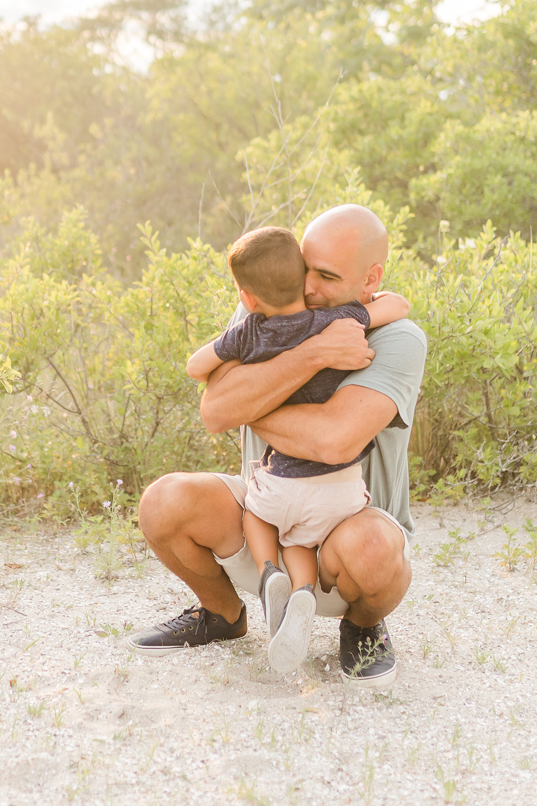 A happy father squats and hugs his toddler son on a sandy beach trail