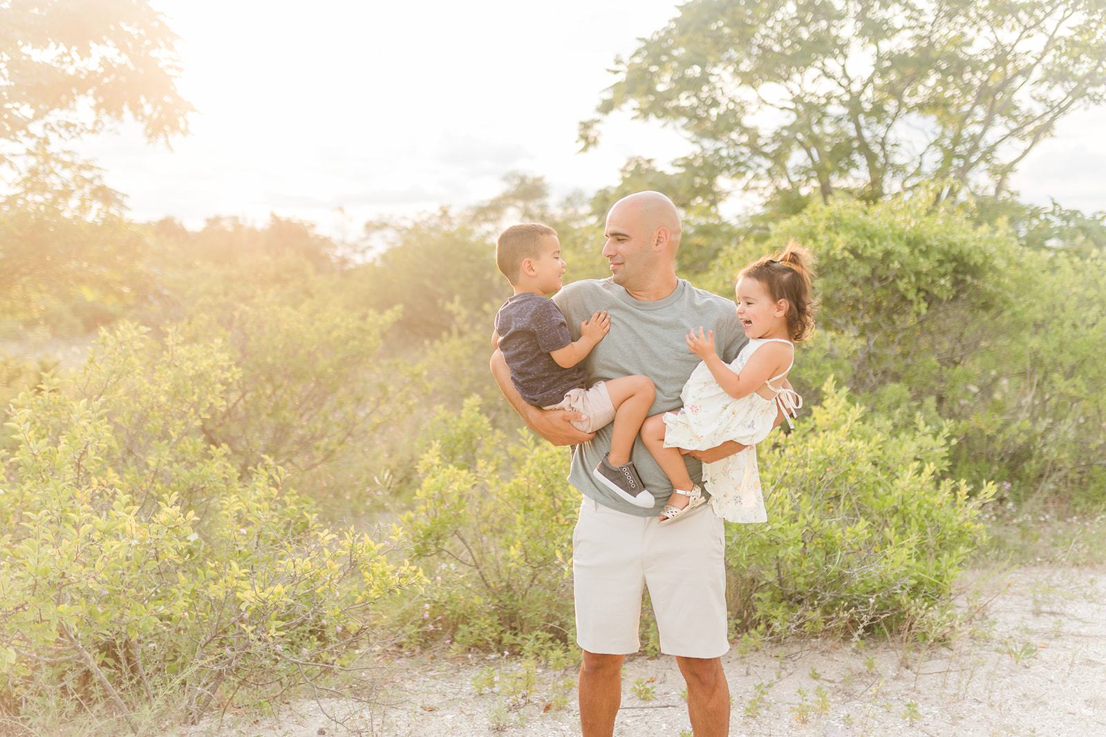 A father in a grey shirt stands in a beach trail holding his toddler children in each arm after visiting Preschools in Trumbull CT