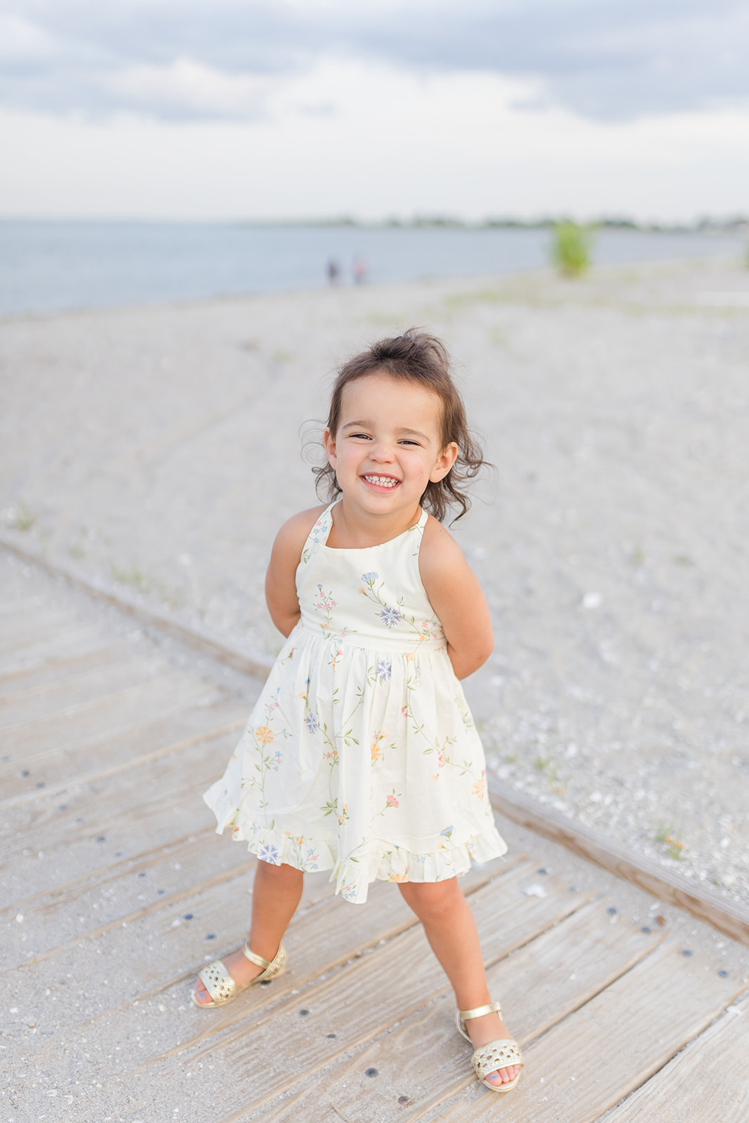 A toddler girl in a white dress stands on a beach boardwalk smiling after visiting Preschools in Trumbull CT