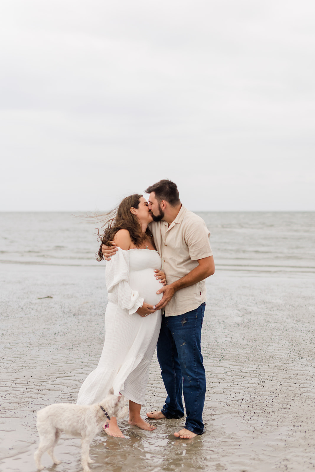 A mom to be kisses her husband while standing on the beach with hands on the bump as their small dog walks around them