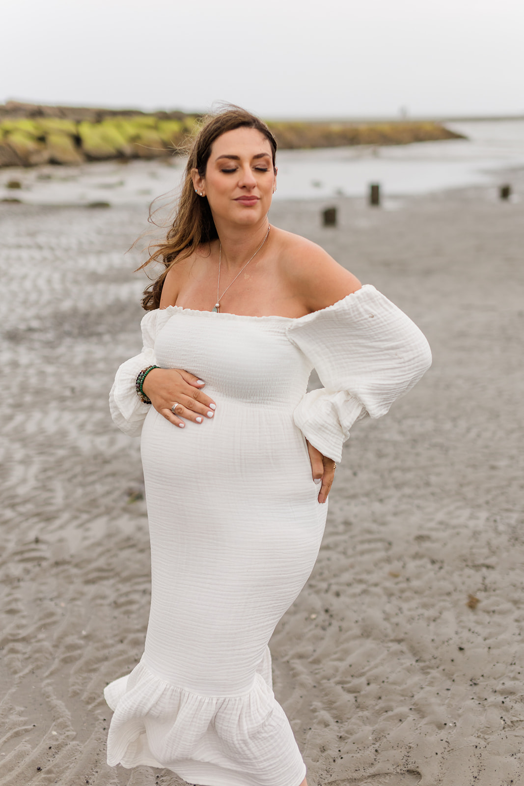A mom to be stands on a beach in a white maternity dress on a windy day after visiting Sabita Holistic Center