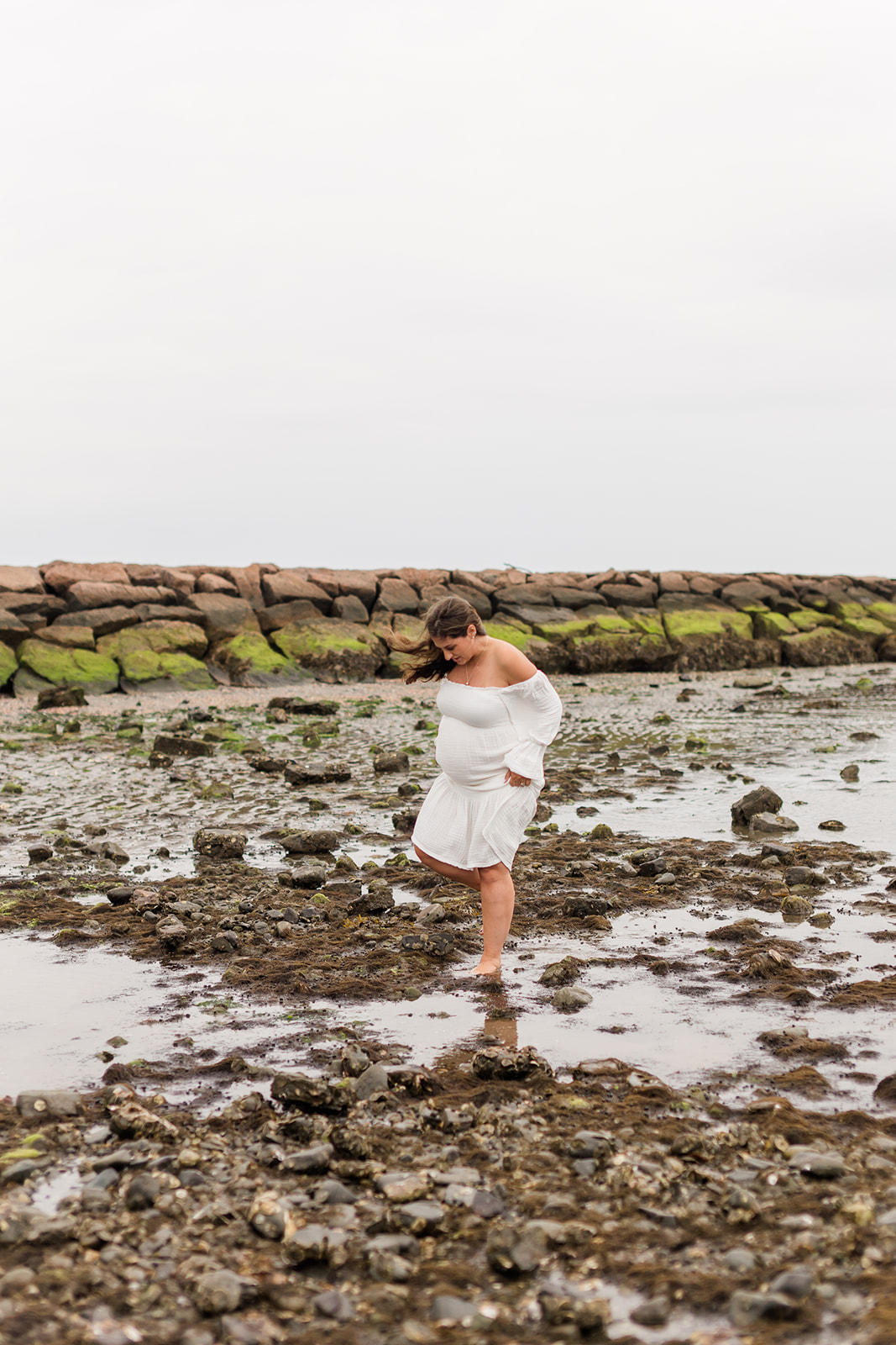A mom to be in a white maternity gown walks through a rocky beach after visiting Sabita Holistic Center