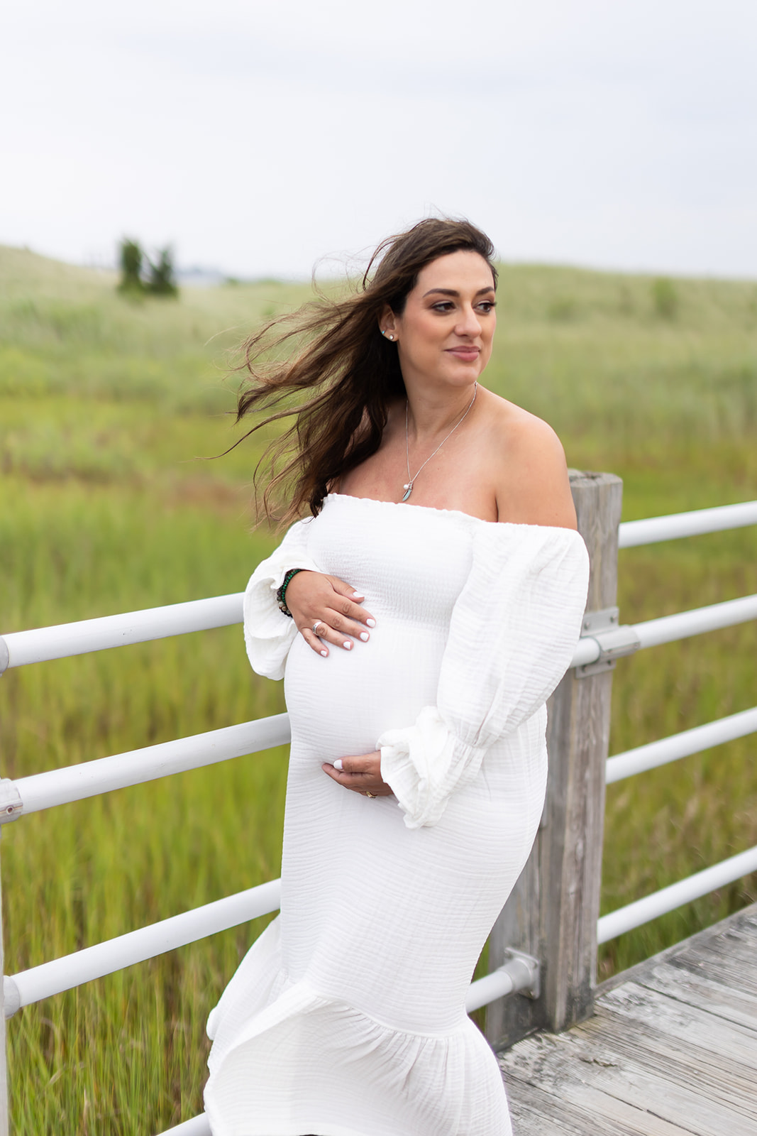 A pregnant woman in a white maternity gown smiles over her shoulder while walking on a windy boardwalk