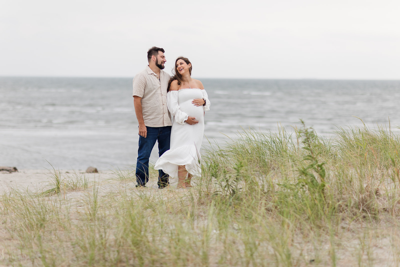 A mother to be in a white dress stands on a windy beach with her husband while laughing after visiting Yale Labor and Delivery
