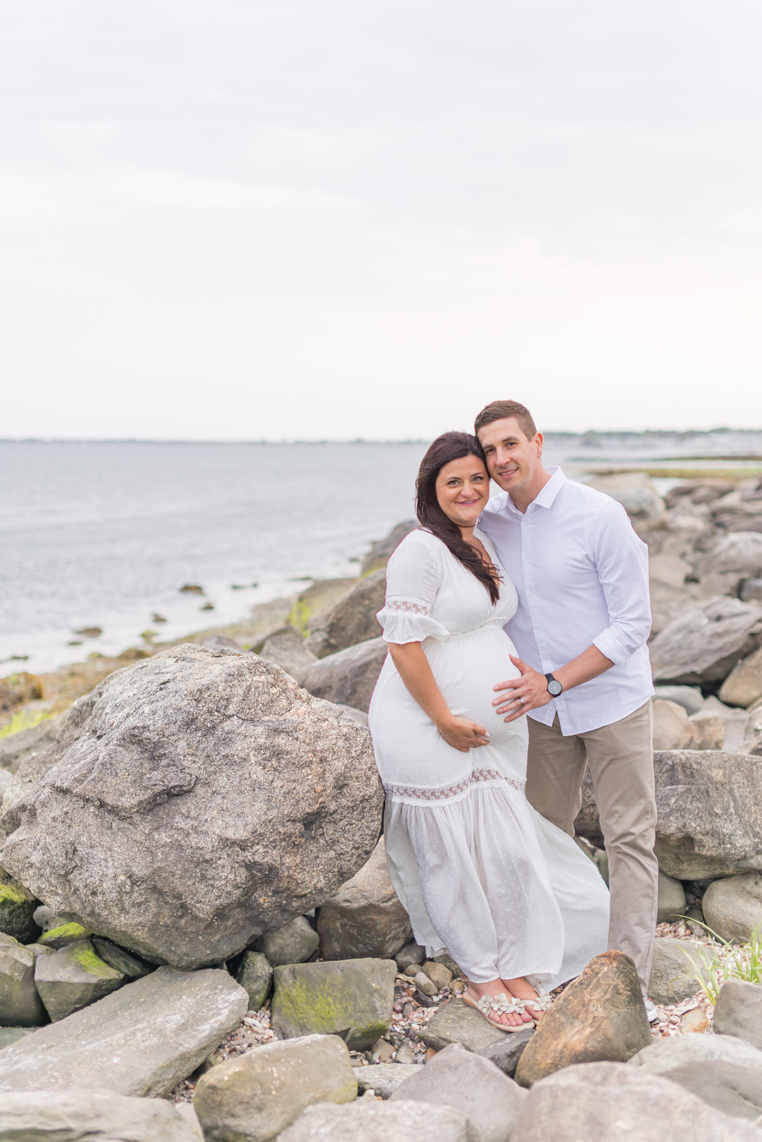 A mom to be and dad to be lean on a large rock on a beach with hands on the bump