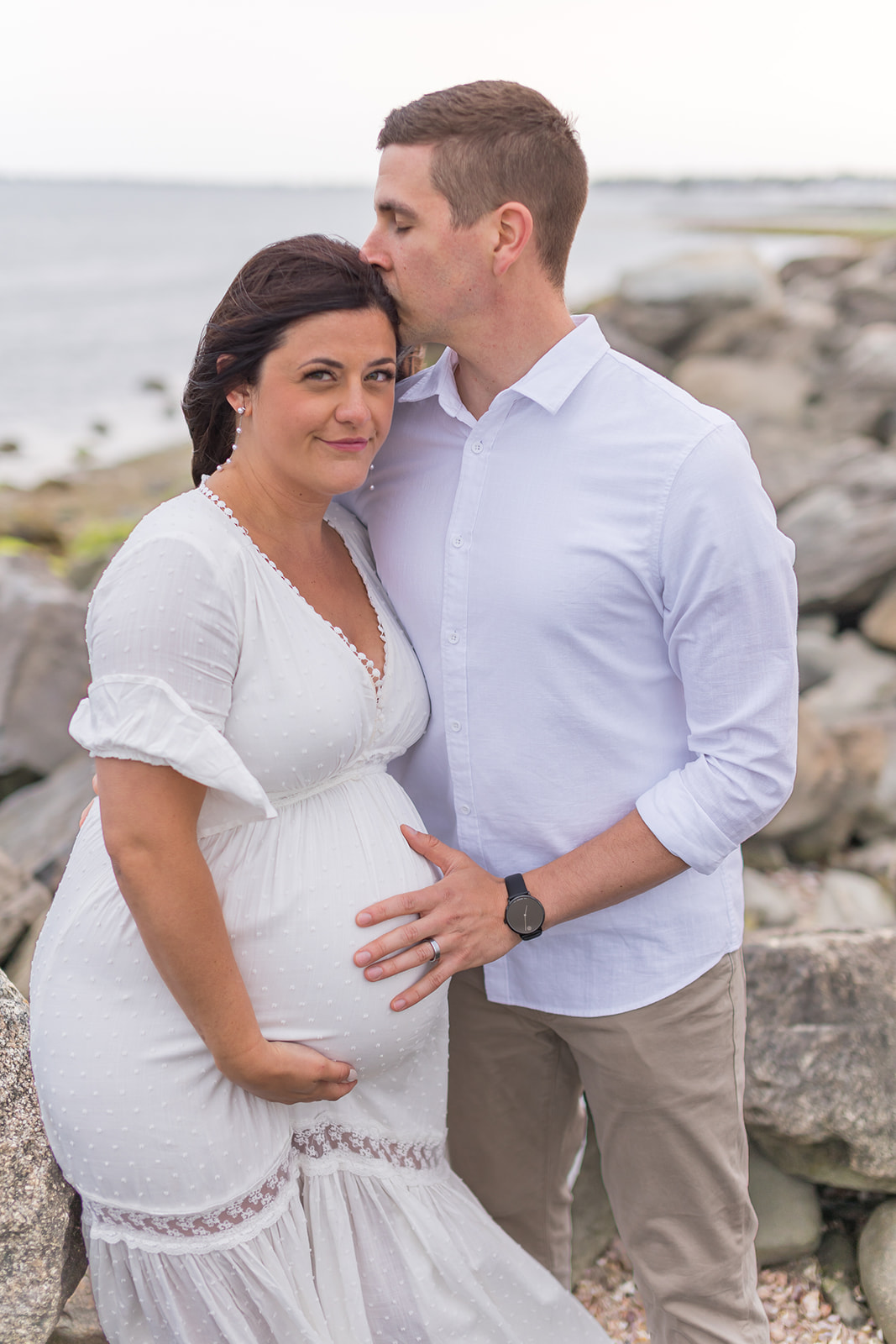 A mom to be leans into her husband in a white maternity dress on a rocky beach thanks to Yale Midwives