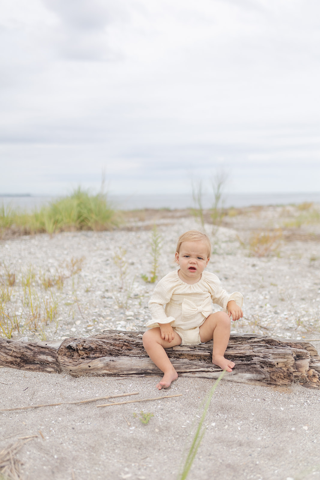 A toddler girl plays while sitting on a piece of driftwood on a beach