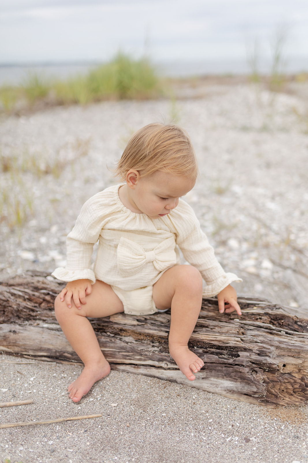 A toddler girl in a yellow onesie sits on a piece of driftwood on a beach before visiting Dwell New Haven